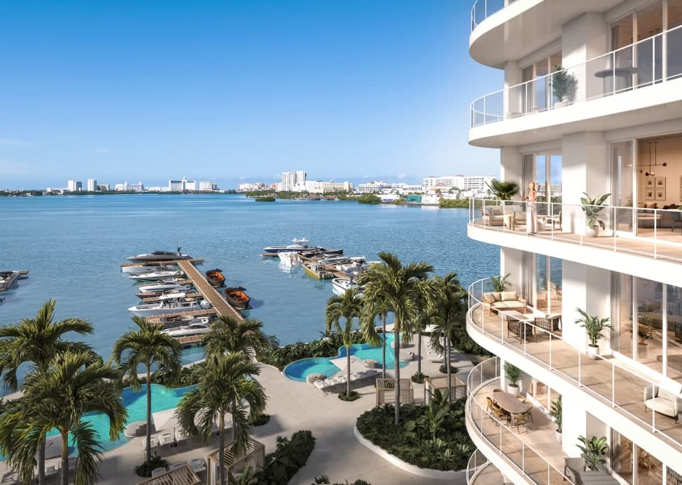View from a luxury waterfront residence showing curved balconies with outdoor seating overlooking a marina with boats docked on calm blue water, palm trees and a landscaped pool deck below, and a city skyline across the bay.