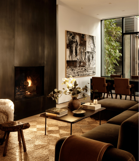 A warm and sophisticated living space featuring a modern fireplace with a sleek black surround. The room is furnished with a plush dark-toned sofa, a wooden coffee table adorned with books and decorative items, and a textured area rug. A large abstract artwork hangs on the wall, while floor-to-ceiling windows let in natural light and offer a view of lush greenery outside. In the background, a dining area with stylish chairs adds to the elegant ambiance.