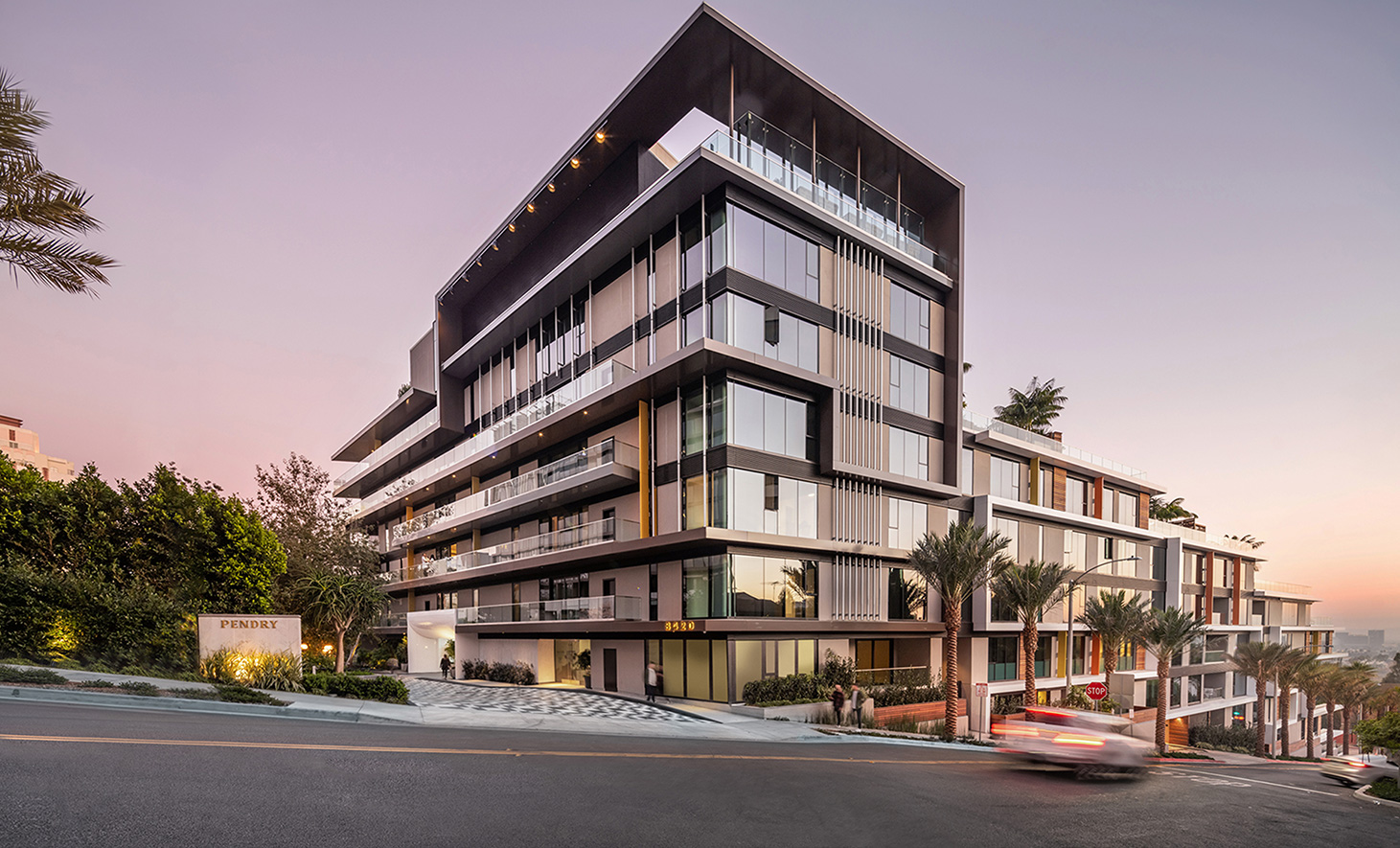 A rendering of the Pendry Residences in West Hollywood, CA. The building is a modern, multi-story building with sleek architectural design, featuring large glass windows, clean lines, and spacious balconies. The structure is set on a hillside with palm trees and greenery surrounding it. The sky is a soft pastel hue, suggesting early evening or sunset. A car with motion blur drives by on the street, adding a dynamic element to the scene.